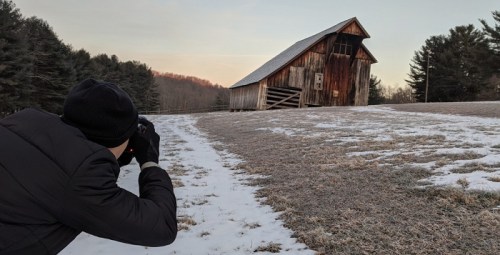 A person in a black coat photographs a leaning wooden barn on a snowy field surrounded by trees.