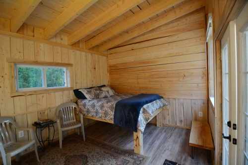 Cozy wooden interior of a small cabin featuring a raised bed, chairs, and a bench, with natural light from windows.