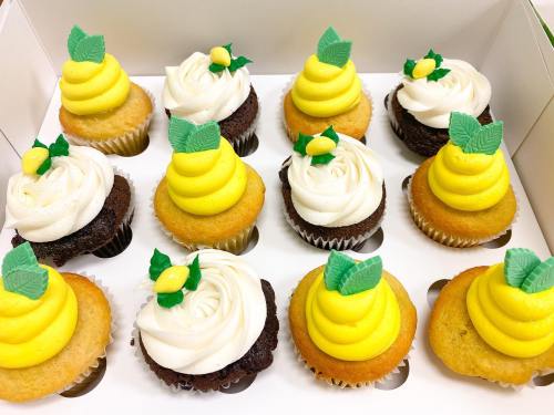 A box of twelve cupcakes with yellow and white frosting, topped with green leaves and lemon decorations.