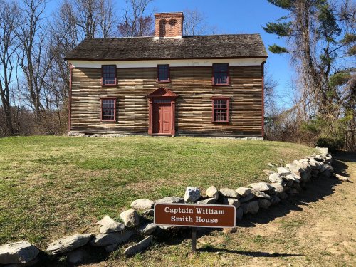 Historic wooden house with a red door and windows, surrounded by grass and trees, featuring a sign that reads "Captain William Smith House."