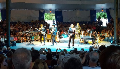 A band performs on stage in a tent, with a large audience watching from below. Instruments and equipment are visible.