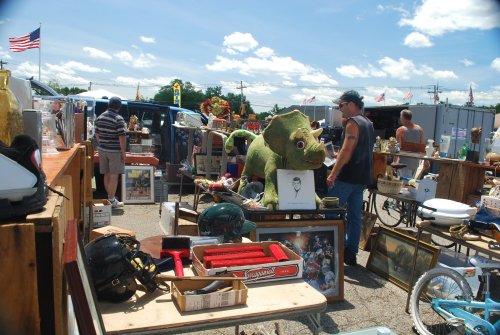 A bustling flea market with various antiques, a large dinosaur statue, and shoppers browsing among the stalls.
