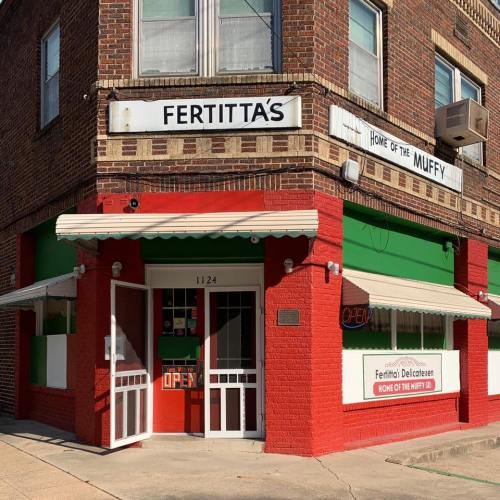 Exterior of Fertitta's Delicatessen, featuring red brick, green accents, and an "OPEN" sign. Home of the Muffy.