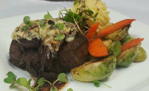 A gourmet dish featuring a beef filet topped with cheese, served with roasted vegetables and microgreens on a white plate.