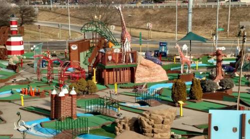 Colorful mini-golf course featuring animal sculptures, bridges, and water features, set against a highway backdrop.