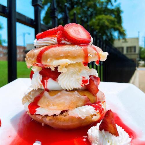 A towering dessert of donuts layered with whipped cream, strawberries, and strawberry sauce on a white plate.