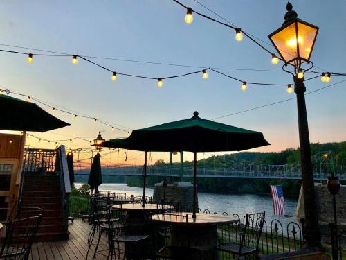 A riverside patio at sunset, with string lights, tables, and an American flag in the background.