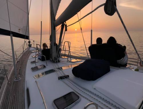 A couple sits on a sailboat, watching a sunset over calm waters, with sails gently billowing in the breeze.