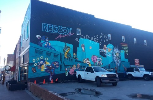 Colorful mural on a building featuring various cartoon characters and retro gaming elements, with parked trucks in front.