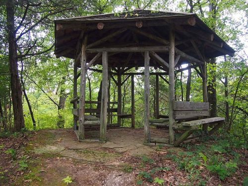 A wooden gazebo surrounded by trees, featuring benches and a stone floor, set in a natural, green landscape.