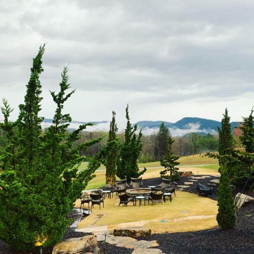 A scenic outdoor area with tables and chairs surrounded by tall trees, overlooking misty mountains under a cloudy sky.