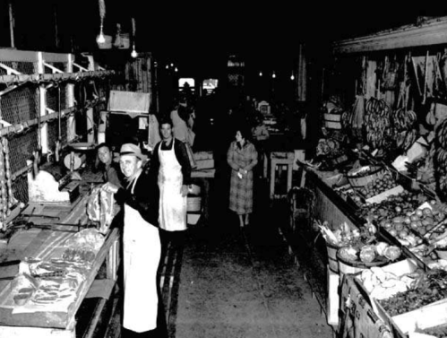 A vintage black-and-white photo of a bustling market with vendors, produce, and customers in a narrow aisle.