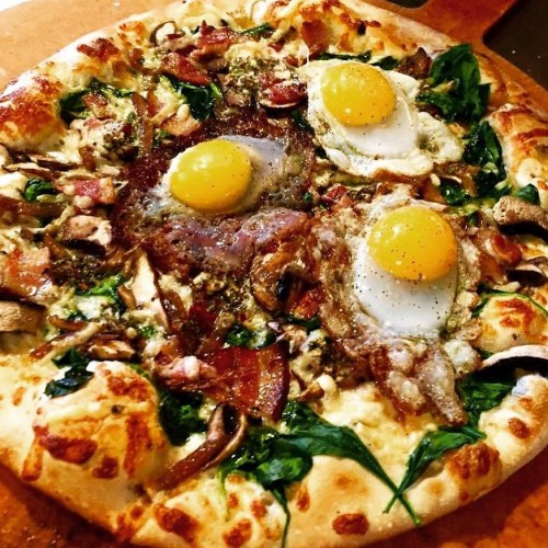 A delicious pizza topped with spinach, mushrooms, bacon, and three sunny-side-up eggs.