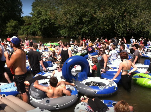 A crowded river scene with people relaxing on colorful inner tubes under a sunny sky surrounded by trees.