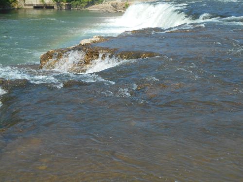A flowing river with cascading water over rocky ledges, surrounded by greenery and a clear blue sky.