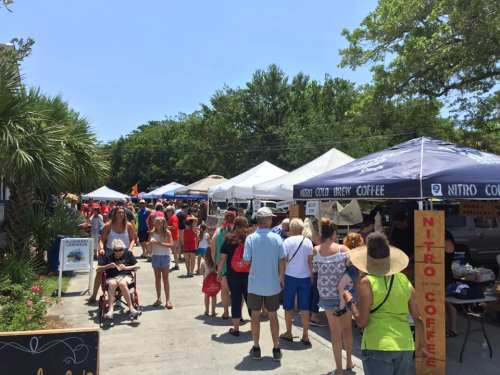 A bustling outdoor market scene with people browsing stalls and tents under sunny skies and trees.