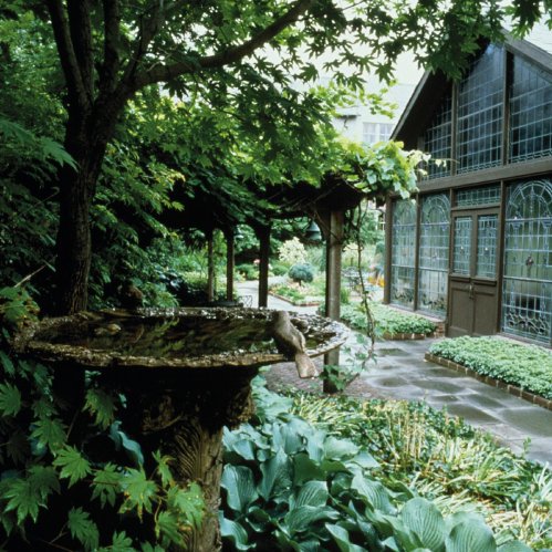 Lush garden scene featuring a birdbath, greenery, and a charming glass-paneled building in the background.