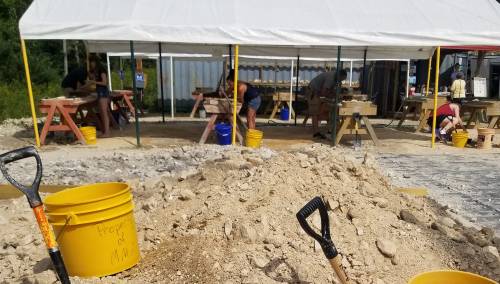 People working under a tent at a construction site, with tools and piles of dirt in the foreground.