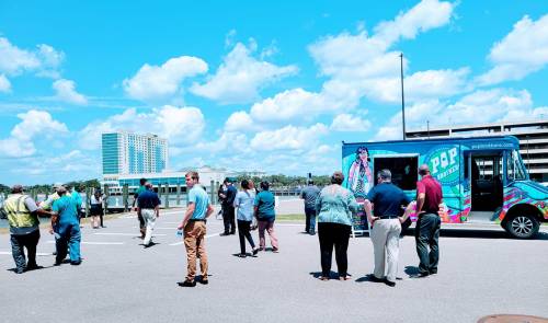 A group of people gathers around a colorful food truck on a sunny day with blue skies and fluffy clouds.