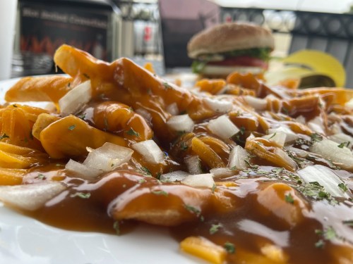 A close-up of a plate of poutine topped with gravy, cheese curds, and chopped onions, with a burger in the background.