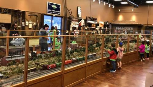 Children and adults explore a model train display in a museum, surrounded by detailed landscapes and miniature buildings.