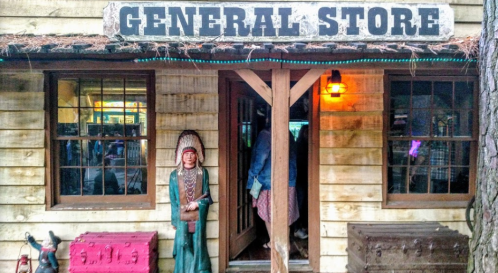 A rustic general store with wooden siding, a sign above, and a carved figure of a Native American outside.
