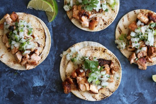 Four tacos filled with seasoned chicken, diced onions, and cilantro, served with lime wedges on a blue background.