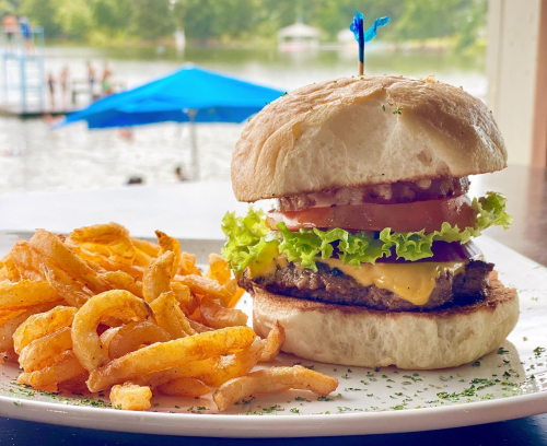 A juicy cheeseburger with lettuce, tomato, and onion, served with curly fries, against a scenic lake backdrop.