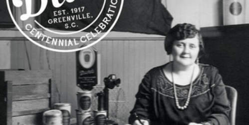 Black and white photo of a woman seated at a desk with vintage items, celebrating a centennial event.