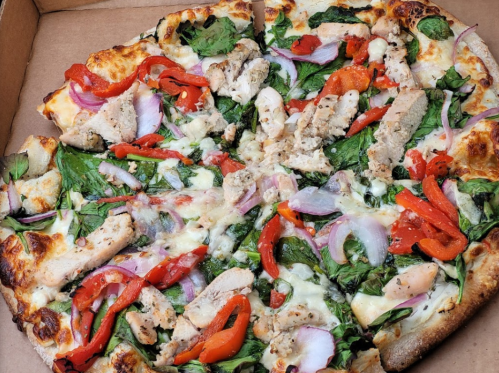 A pizza topped with spinach, red peppers, onions, and grilled chicken in a pizza box.