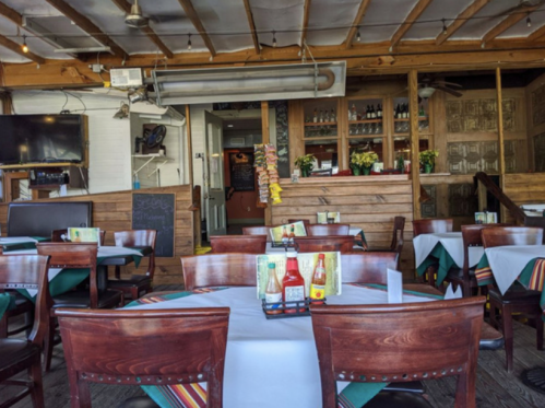 Interior of a restaurant featuring wooden tables, chairs, and a bar area with condiments on the tables.