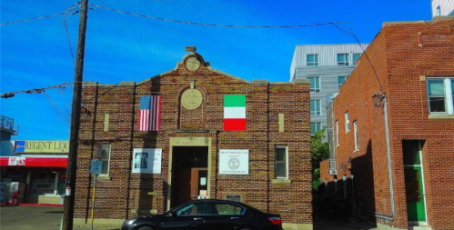Brick building with an American flag and an Italian flag, set against a clear blue sky. Nearby buildings visible.
