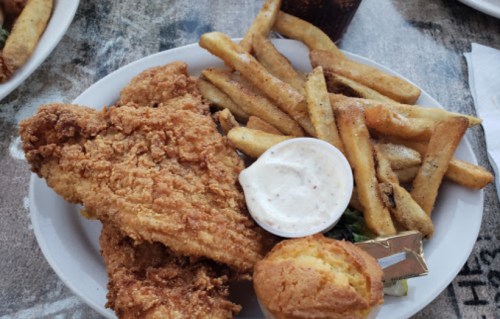 A plate of crispy fried chicken, French fries, a small cornbread muffin, and a cup of dipping sauce.
