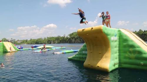 A person jumps off a large inflatable slide into a lake, with others enjoying water activities nearby.