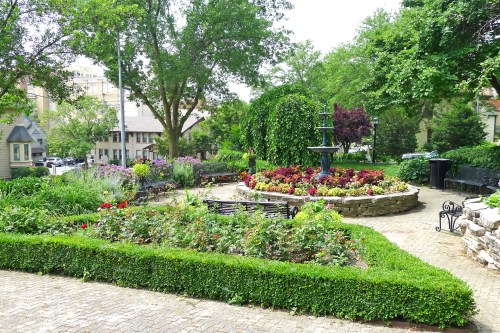 A lush garden with colorful flowers, neatly trimmed hedges, and a stone path, surrounded by trees and benches.