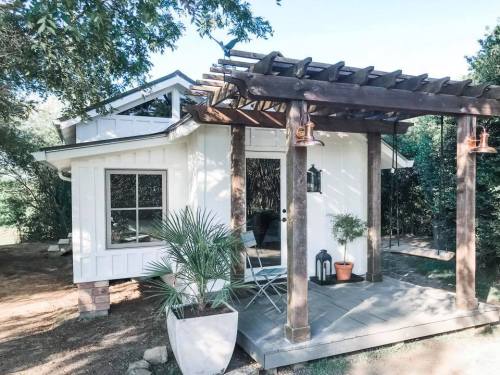 A charming white cottage with a wooden pergola, potted plants, and a small patio area surrounded by greenery.