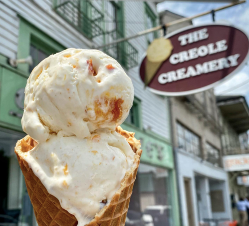 A close-up of a waffle cone filled with two scoops of ice cream, with The Creole Creamery sign in the background.