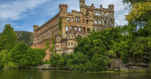 A historic, overgrown castle partially hidden by trees, reflecting in a calm body of water under a blue sky.