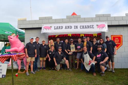 A group of people in matching shirts pose in front of a castle-themed booth with a pig statue, celebrating a bacon event.