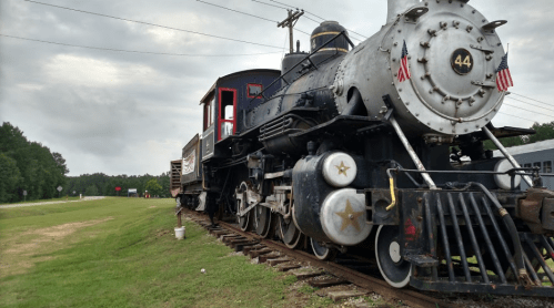 A vintage steam locomotive with American flags parked on a grassy area beside a road under a cloudy sky.