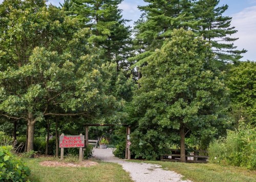 A pathway leads through lush greenery to a sign reading "Nature Explore Classroom" amidst tall trees.