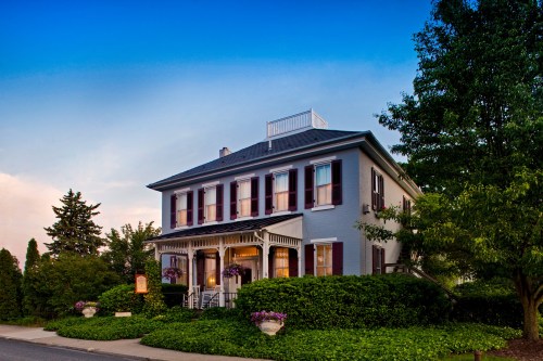 A charming two-story house with a porch, surrounded by greenery and flowers, under a clear blue sky.