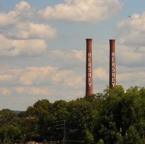Two tall smokestacks with "HERSHEY" written on them, surrounded by trees and a cloudy blue sky.