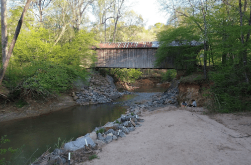 A rustic covered bridge spans a calm river, surrounded by lush greenery and sandy banks.