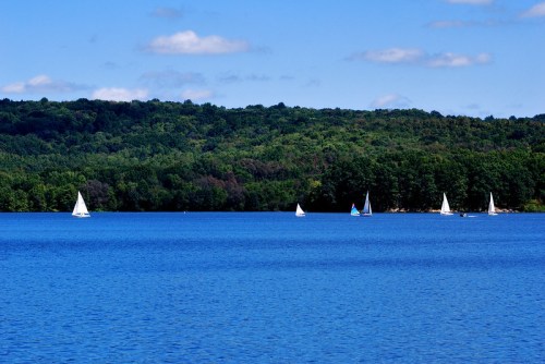 Sailboats glide across a calm blue lake, surrounded by lush green hills under a clear blue sky.