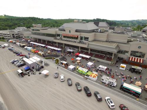 Aerial view of a bustling outdoor event with food trucks and tents near a large building, surrounded by parked cars.