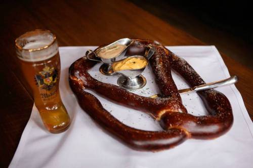 A large pretzel with salt, served with two dips, alongside a glass of beer on a wooden table.