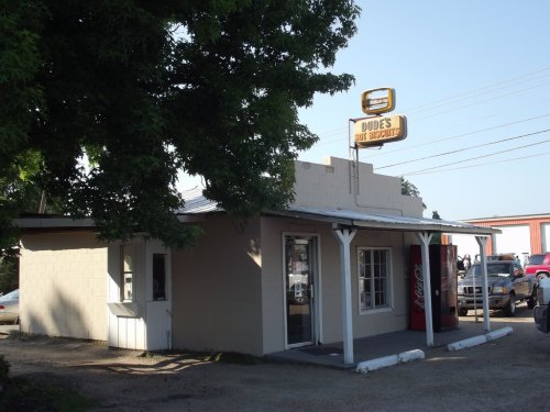 A small, single-story building with a sign reading "Dude's Hot Biscuit," surrounded by trees and parked cars.
