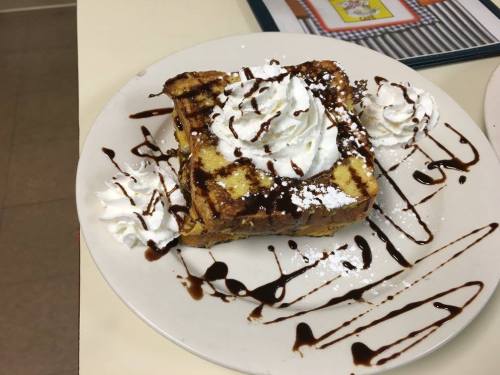 A slice of French toast topped with whipped cream, powdered sugar, and chocolate drizzle, served on a white plate.