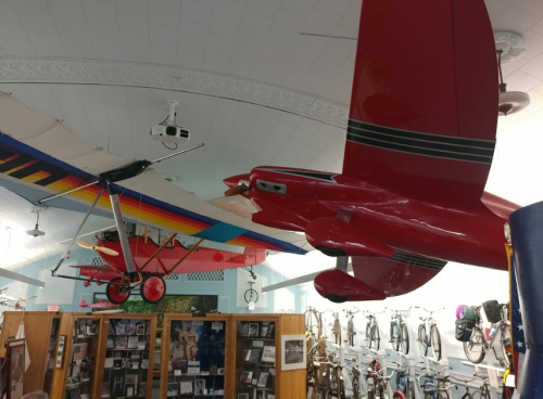 Two model airplanes hang from the ceiling of a museum, with a display of bicycles in the background.